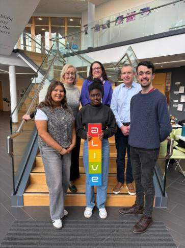 6 people standing in front of stairs, one of them holds the ALIVE sign.