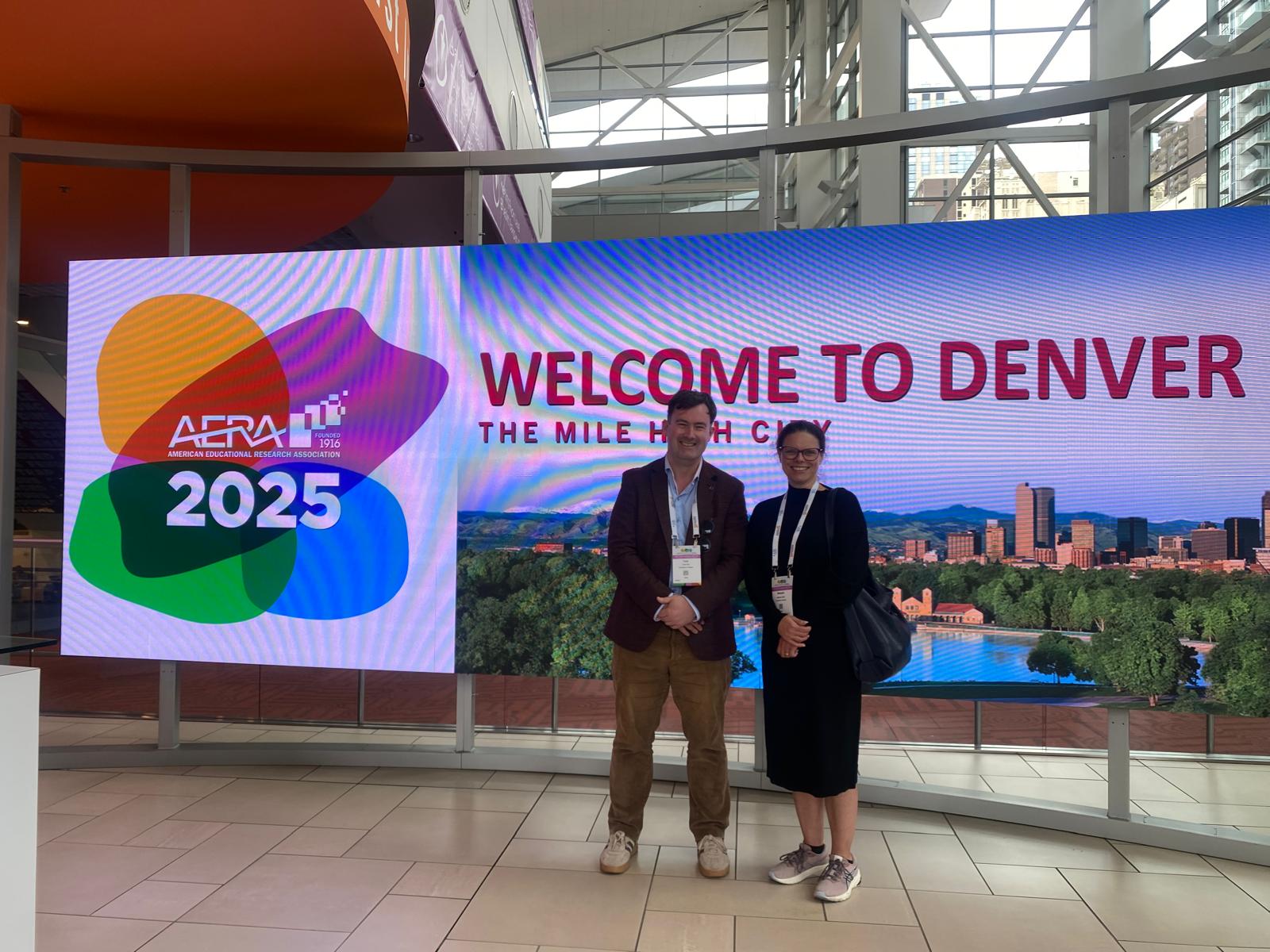 Prof Tony Hall and Dr Manuela Heinz standing in front of a video wall displaying the AERA logo