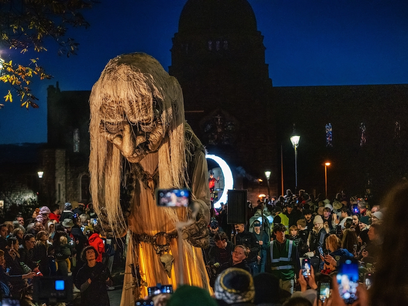 Macnas Parade in Galway featuring a giant puppet. Crowds take pictures as the puppet passes the bridge in front of the cathedral.